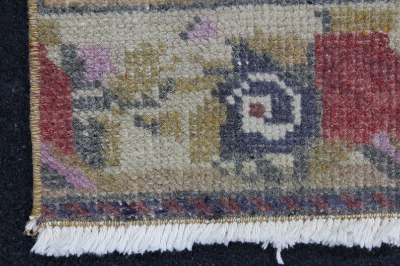 May include: Close-up of a section of a small, hand-woven rug. The rug features a complex pattern with colors including red, blue, yellow, and purple. The rug has a fringe along one edge.