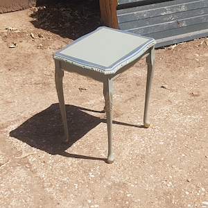 Op de afbeelding: Een kleine, lichtblauwe bijzettafel met een decoratief, gesneden ontwerp op de poten en een gouden rand rond de bovenkant. De tafel staat op een grind oppervlak.