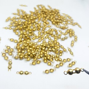 May include: A large quantity of gold-coloured metal beads with a loop on each side. The beads are scattered on a white surface.