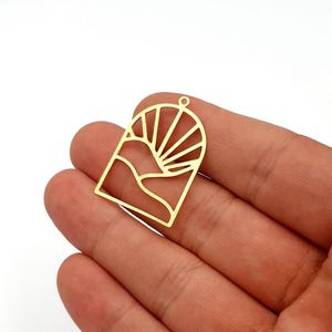 Könnte beinhalten: Goldfarbener Metallanhänger mit einem ausgeschnittenen Design einer Berglandschaft und Sonnenstrahlen. Der Anhänger hat oben eine kleine Schlaufe zum Befestigen an einer Kette. Das Design ist geometrisch und minimalistisch.