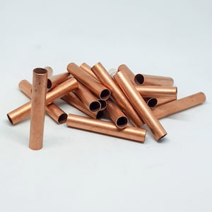 May include: A collection of copper tubes, cylindrical in shape, scattered on a white surface. The tubes have a shiny, metallic finish and appear to be hollow. The ends of the tubes are visible, showing the open interior. The copper color is consistent throughout.