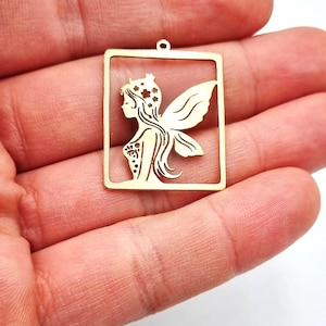 Può includere: Ciondolo color oro con un design a forma di fata all'interno di una cornice quadrata. La fata ha lunghi capelli, una corona e ali. Il ciondolo ha un piccolo anello nella parte superiore per appenderlo.
