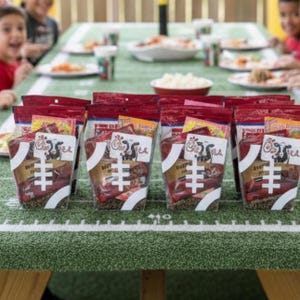 May include: A selection of clear plastic food bags with red tops and white football graphics are displayed on a green table with white lines, mimicking a football field. Children are seated around the table, ready to eat.