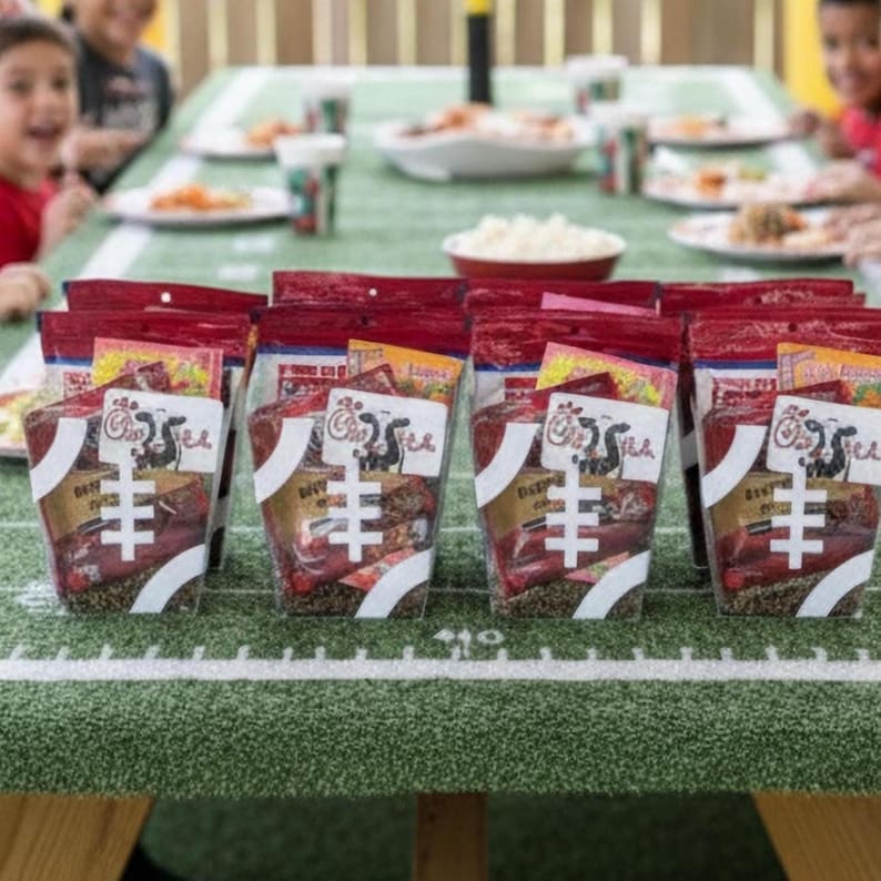 May include: A selection of clear plastic food bags with red tops and white football graphics are displayed on a green table with white lines, mimicking a football field. Children are seated around the table, ready to eat.
