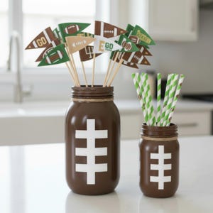 May include: Two brown mason jars decorated with white football designs. One jar holds football-themed pennant flags on wooden sticks, the other holds green and white striped straws. The jars are on a white surface.