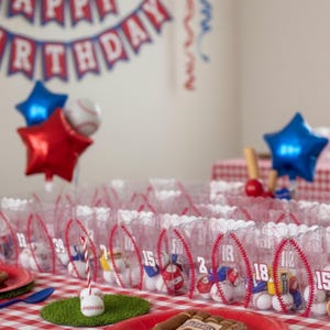 May include: A baseball-themed birthday party setup with clear favor bags decorated with red stitching and numbers. The bags are filled with baseballs and small toys. Red and blue star-shaped balloons and a banner that says "Happy Birthday" complete the festive scene.
