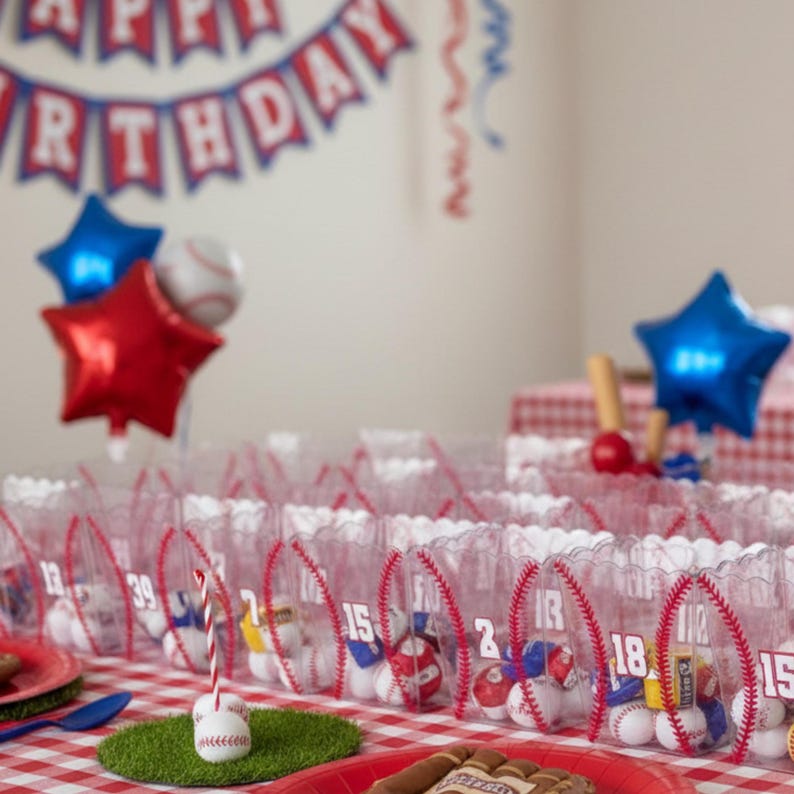 May include: A baseball-themed birthday party setup with clear favor bags decorated with red stitching and numbers. The bags are filled with baseballs and small toys. Red and blue star-shaped balloons and a banner that says "Happy Birthday" complete the festive scene.