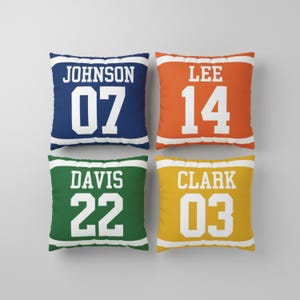 May include: Four square throw pillows featuring a sports jersey design with white horizontal stripes at the top and bottom. The pillows are navy blue with JOHNSON 07, orange with LEE 14, green with DAVIS 22, and yellow with CLARK 03 in white text.