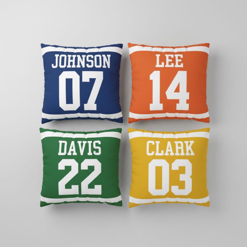 May include: Four square throw pillows featuring a sports jersey design with white horizontal stripes at the top and bottom. The pillows are navy blue with JOHNSON 07, orange with LEE 14, green with DAVIS 22, and yellow with CLARK 03 in white text.
