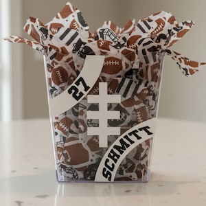 May include: A transparent plastic container filled with football-themed tissue paper. The container has a white banner with the number "27" and the name "SCHMITT". The tissue paper features brown footballs and black football helmets.