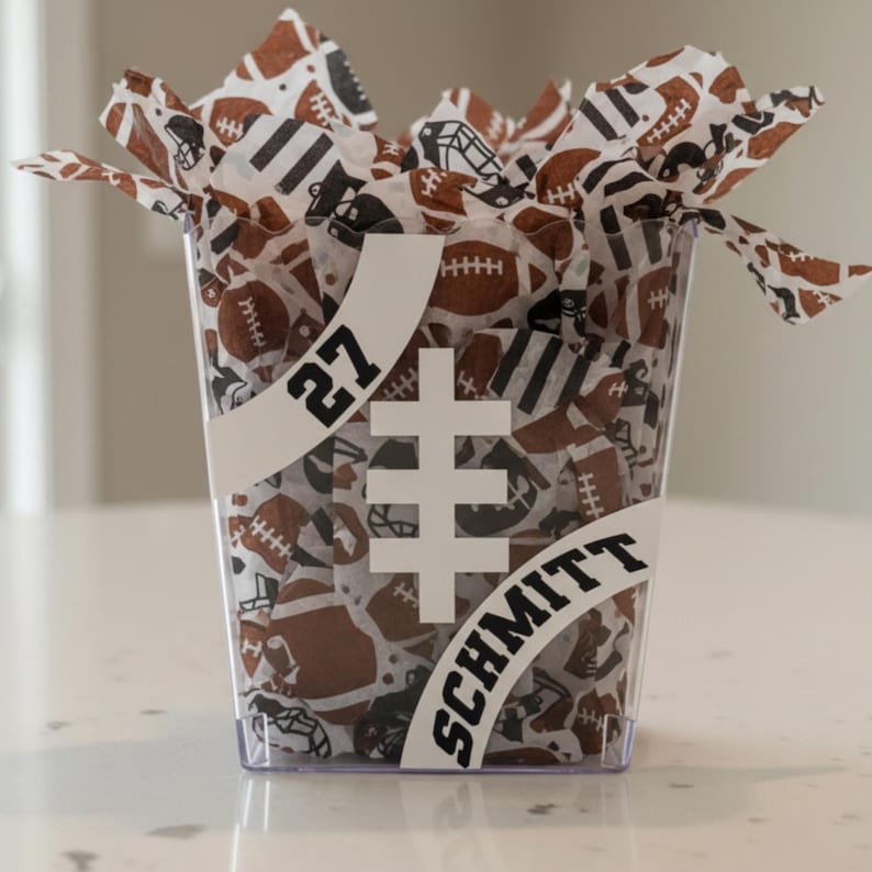 May include: A transparent plastic container filled with football-themed tissue paper. The container has a white banner with the number "27" and the name "SCHMITT". The tissue paper features brown footballs and black football helmets.