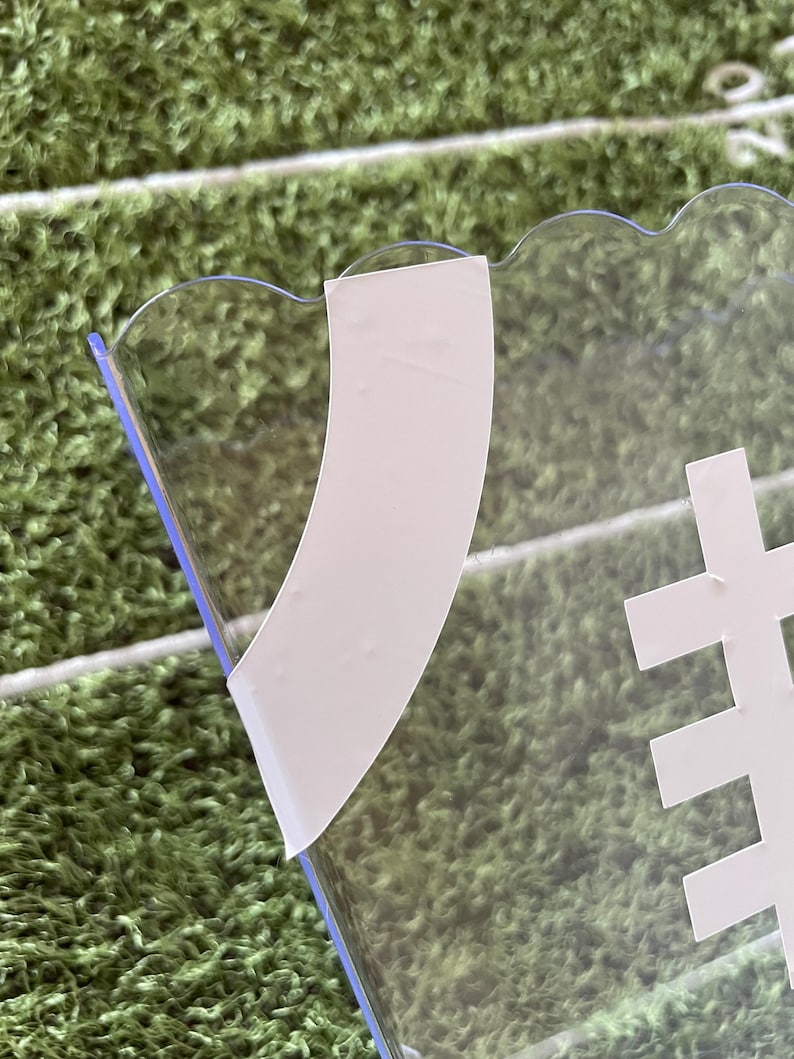 May include: A clear, football-shaped serving dish with a scalloped edge. White tape is applied to the clear plastic. A white football graphic is visible on the dish. The background is a green field.