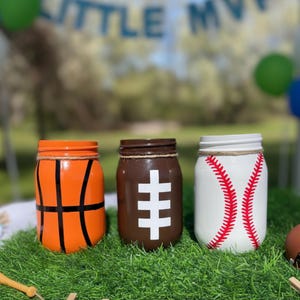 May include: Three painted mason jars decorated as sports balls: a basketball, a football, and a baseball. The basketball jar is orange with black lines. The football jar is brown with a white cross. The baseball jar is white with red stitching.
