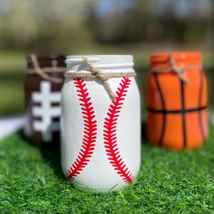 May include: Three painted mason jars decorated to resemble sports balls. The front jar is white with red baseball stitching. The other jars are painted to look like a football and a basketball. Each jar has a twine accent around the rim.