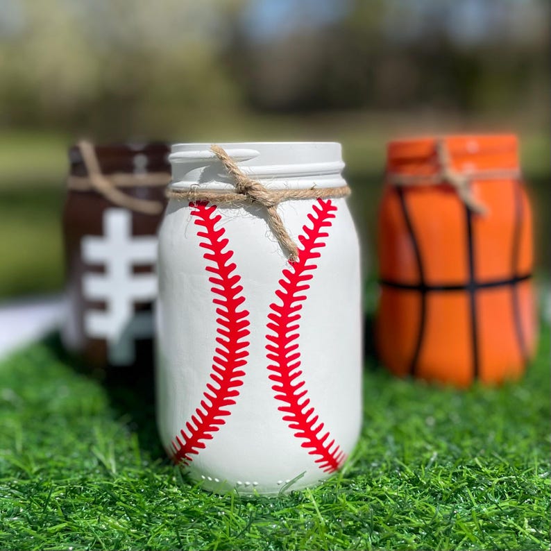May include: Three painted mason jars decorated to resemble sports balls. The front jar is white with red baseball stitching. The other jars are painted to look like a football and a basketball. Each jar has a twine accent around the rim.
