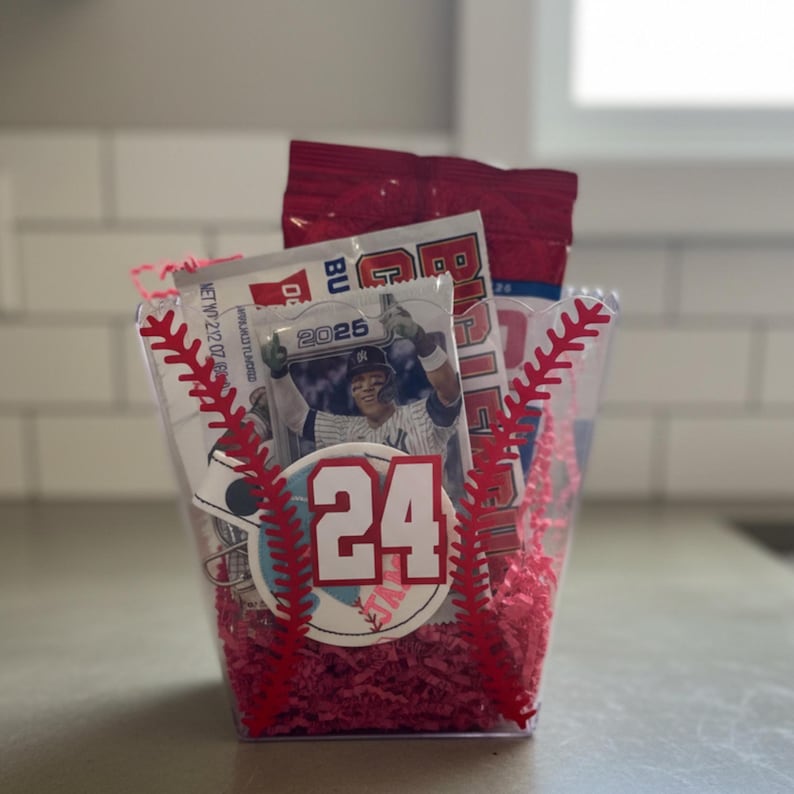 May include: A clear plastic container filled with red paper shreds, a baseball card, and a red snack bag. The container is decorated with red baseball stitching and a baseball-themed sticker with the number 24.