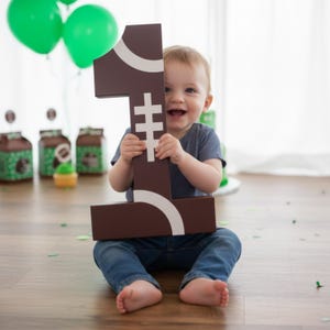 May include: A smiling baby sits on a wooden floor, holding a large brown number one. The number has white accents and resembles a football. Green balloons and party favors are in the background, suggesting a first birthday celebration.