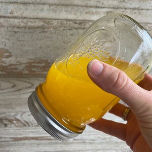 May include: A glass jar filled with a bright yellow liquid, likely honey or a similar viscous substance. The jar has a silver lid and is being held by a hand.