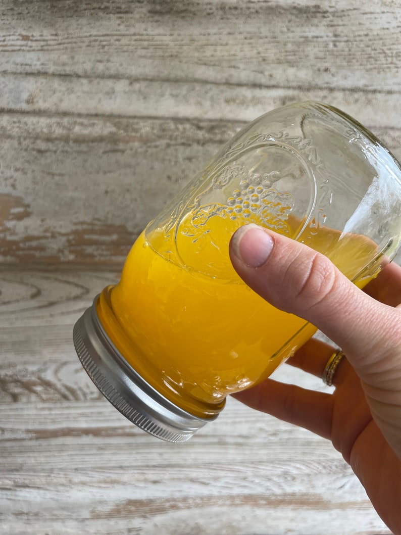 May include: A glass jar filled with a bright yellow liquid, likely honey or a similar viscous substance. The jar has a silver lid and is being held by a hand.