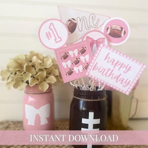 May include: Pink and white printable party decorations for a football themed first birthday party. The decorations include a banner that says "one", a circle with the number "1" and the text "#1", a circle with a football, a banner with a bow, a banner with footballs and bows, and a banner that says "happy birthday".