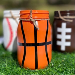 May include: Three painted mason jars decorated as sports balls. The front jar is orange with black lines to resemble a basketball. A baseball jar and a football jar are in the background. Each jar has twine around the rim.