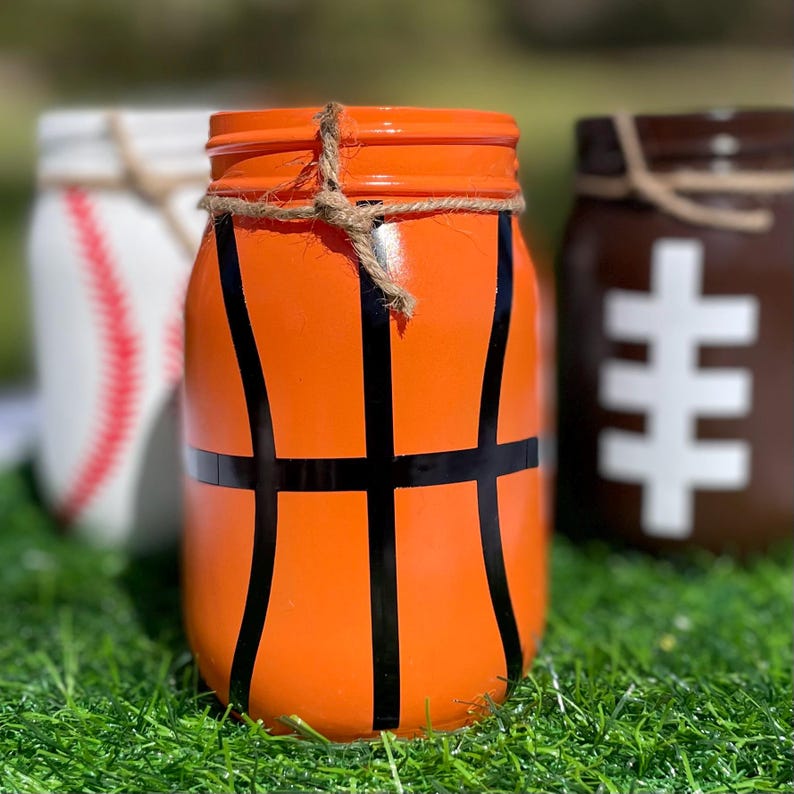 May include: Three painted mason jars decorated as sports balls. The front jar is orange with black lines to resemble a basketball. A baseball jar and a football jar are in the background. Each jar has twine around the rim.