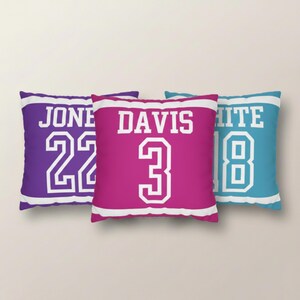 May include: Three square throw pillows featuring a sports jersey design. The left pillow is purple with white text JONES and number 22. The center pillow is pink with white text DAVIS and number 3. The right pillow is blue with white text WHITE and number 18.