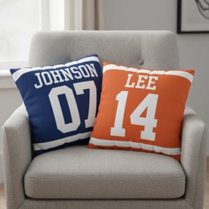 May include: Two square throw pillows featuring a sports jersey design. The left pillow is navy blue with white stripes, the name JOHNSON, and the number 07. The right pillow is orange with white stripes, the name LEE, and the number 14.