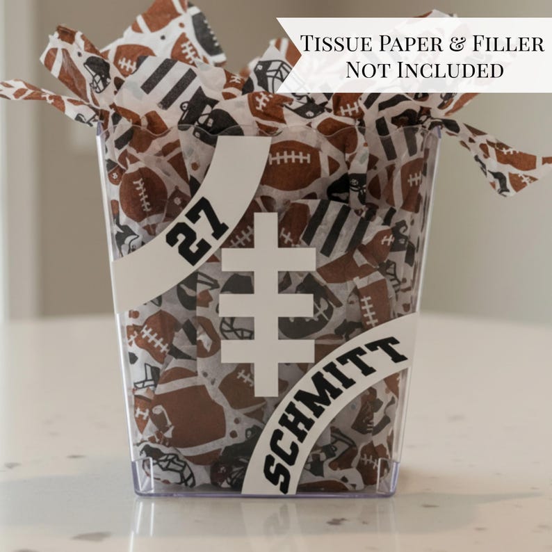 May include: Clear plastic container filled with football-themed tissue paper. The container has a white football graphic, the name "Schmitt" and the number "27" in black. The tissue paper features brown footballs and helmets.