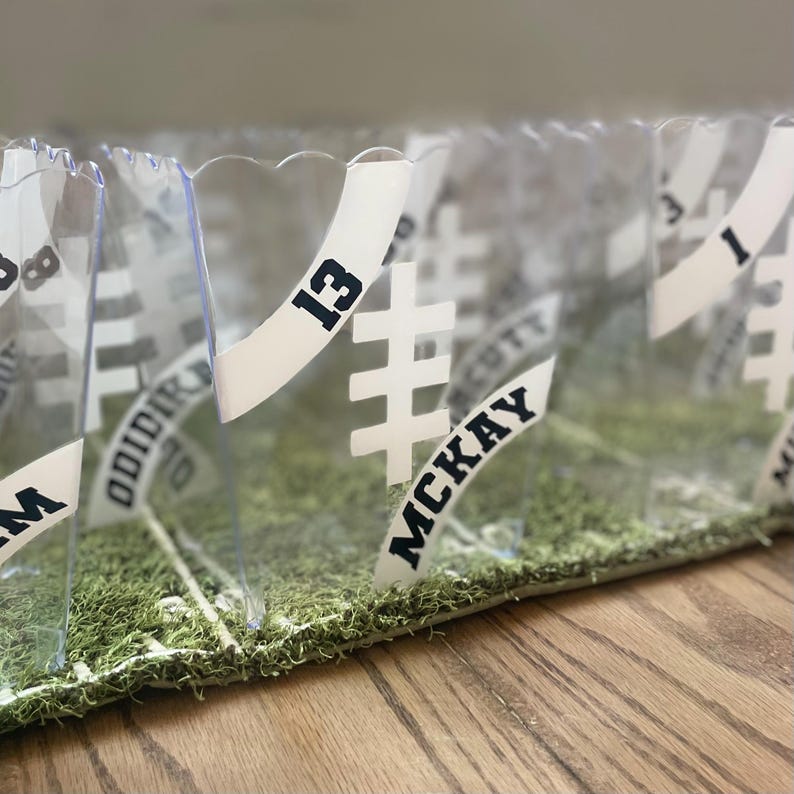 May include: Clear plastic containers with white labels bearing black numbers and names, set on green moss. The labels have a football field design. The number "13" is clearly visible on one label.