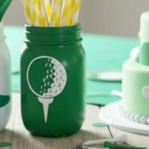 May include: A green mason jar with a white golf ball and tee design. The jar is filled with yellow and white striped straws. A light green cake is visible in the background.