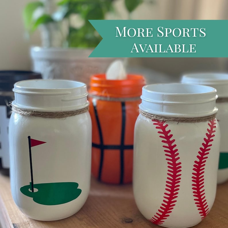 May include: White mason jars decorated with sports themes. One jar has a golf course design, another a baseball, and a third a basketball. The text "MORE SPORTS AVAILABLE" is displayed on a teal banner.
