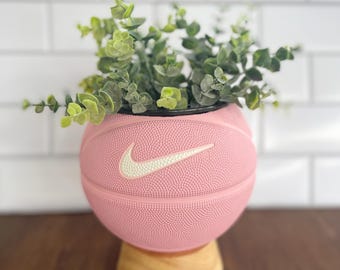 May include: A pink basketball planter with a white Nike logo, filled with green leafy plants. The planter sits on a small, round wooden base. The background is a white tiled wall and a dark wooden surface.