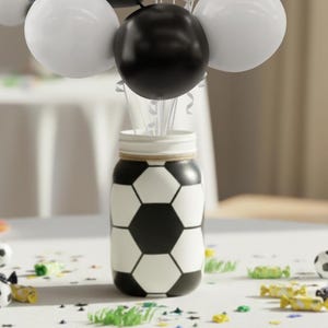 May include: A decorative jar painted with a soccer ball design, holding white and black balloons. The jar is on a white surface with scattered confetti and miniature soccer balls, creating a festive theme.