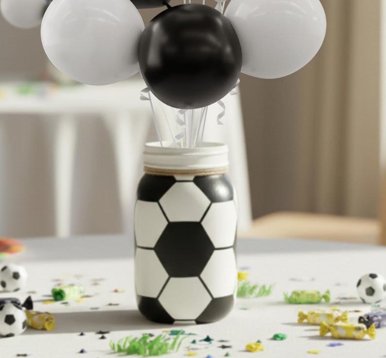 May include: A decorative jar painted with a soccer ball design, holding white and black balloons. The jar is on a white surface with scattered confetti and miniature soccer balls, creating a festive theme.
