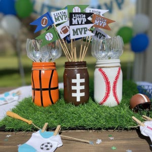 May include: Three mason jars decorated as a basketball, football, and baseball. Each jar holds utensils and is adorned with pennant flags that read "Little MVP." A miniature football helmet and a baseball bat are also present.