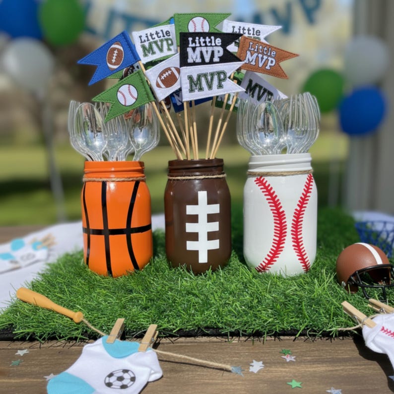 May include: Three mason jars decorated as a basketball, football, and baseball. Each jar holds utensils and is adorned with pennant flags that read "Little MVP." A miniature football helmet and a baseball bat are also present.