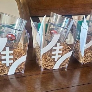 May include: Clear glass tumblers with football-themed graphics are displayed in decorative boxes. The boxes have white accents and are filled with shredded paper. The tumblers have a football graphic and the text "Thank You Co. 2025".