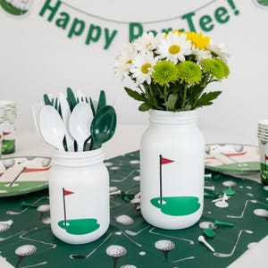 May include: White mason jars decorated with a golf course design, one filled with cutlery and the other with white, yellow, and green flowers. The jars sit on a green golf-themed tablecloth, with a banner that reads "Happy Par-Tee!"