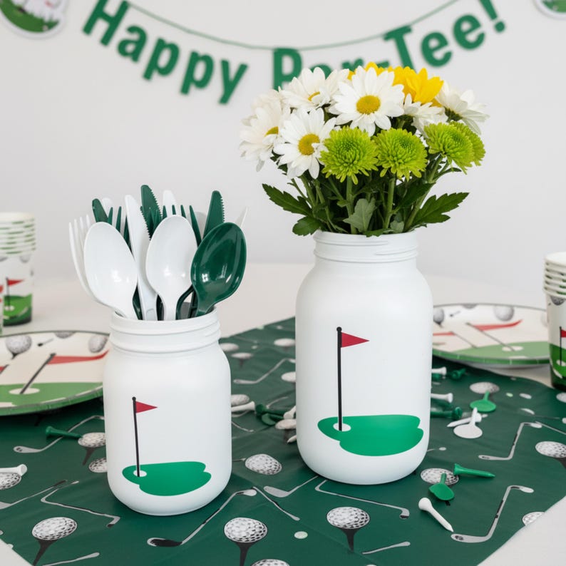 May include: White mason jars decorated with a golf course design, one filled with cutlery and the other with white, yellow, and green flowers. The jars sit on a green golf-themed tablecloth, with a banner that reads "Happy Par-Tee!"