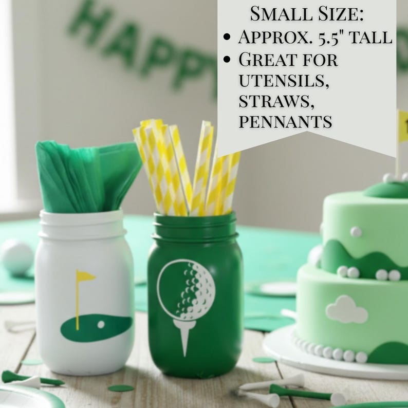 May include: Two mason jars, one white and one green, decorated with golf-themed graphics. The white jar has a golf flag design, while the green jar features a golf ball on a tee. The jars are filled with green napkins and yellow striped straws. A golf-themed cake is in the background.
