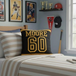May include: A square decorative throw pillow featuring a black background with yellow text that reads MOORE and the number 60 in a bold athletic font. The pillow has yellow horizontal stripes along the top and bottom edges of the front face.