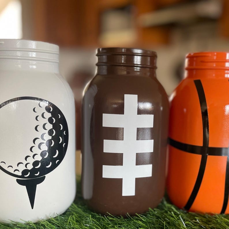 May include: Three painted mason jars with sports themes. One is white with a golf ball design, another is brown with a football design, and the last is orange with a basketball design. The jars are sitting on green turf.