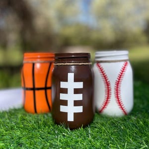 May include: Three painted mason jars decorated as a basketball, football, and baseball. The basketball jar is orange with black lines. The football jar is brown with a white design. The baseball jar is white with red stitching.