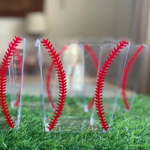 K&ouml;nnte beinhalten: Transparente Plastikbecher mit rotem Baseball-Nahtdesign. Die Becher sind auf einem Bett aus gr&uuml;nem Kunstrasen angeordnet. Das Design &auml;hnelt einem Baseball. Geeignet f&uuml;r eine Party zum Thema Baseball.