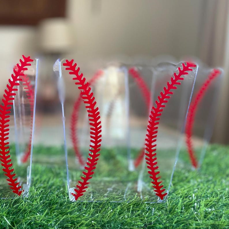 K&ouml;nnte beinhalten: Transparente Plastikbecher mit rotem Baseball-Nahtdesign. Die Becher sind auf einem Bett aus gr&uuml;nem Kunstrasen angeordnet. Das Design &auml;hnelt einem Baseball. Geeignet f&uuml;r eine Party zum Thema Baseball.