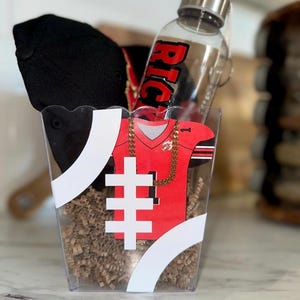 May include: A gift basket containing a black baseball cap, a red and black football jersey cutout, and a clear water bottle with the word "RICH" in red. The basket is decorated with white stripes and filled with brown paper.