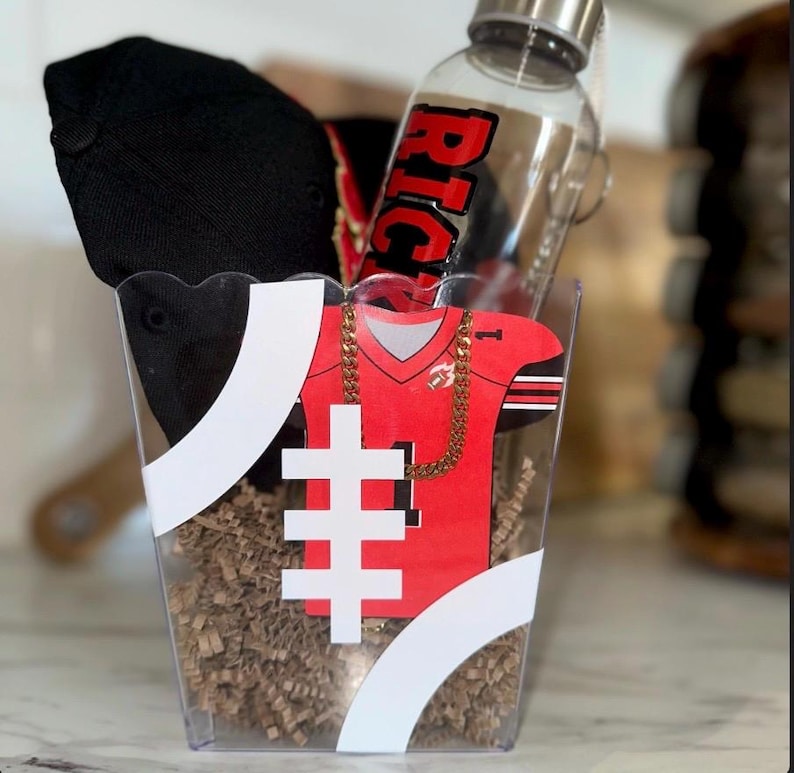 May include: A gift basket containing a black baseball cap, a red and black football jersey cutout, and a clear water bottle with the word "RICH" in red. The basket is decorated with white stripes and filled with brown paper.