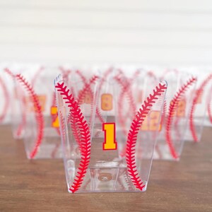 K&ouml;nnte beinhalten: Transparente Plastikbecher mit roten Baseballn&auml;hten und gelben Zahlen. Die Becher sind mit roten N&auml;hten verziert, die einen Baseball nachahmen. Die Zahl "1" ist auf der Vorderseite des Bechers sichtbar. Weitere Becher mit Zahlen sind im Hintergrund.