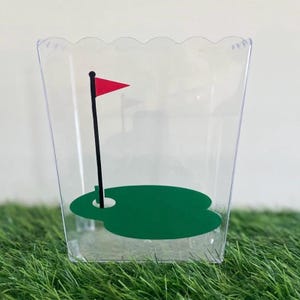 May include: Clear plastic container with a green golf course and a red flag on a black pole. The container has a scalloped edge.